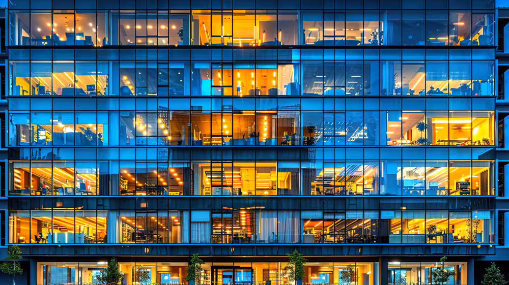Modern office building facade with illuminated glass windows showing interior offices at night.