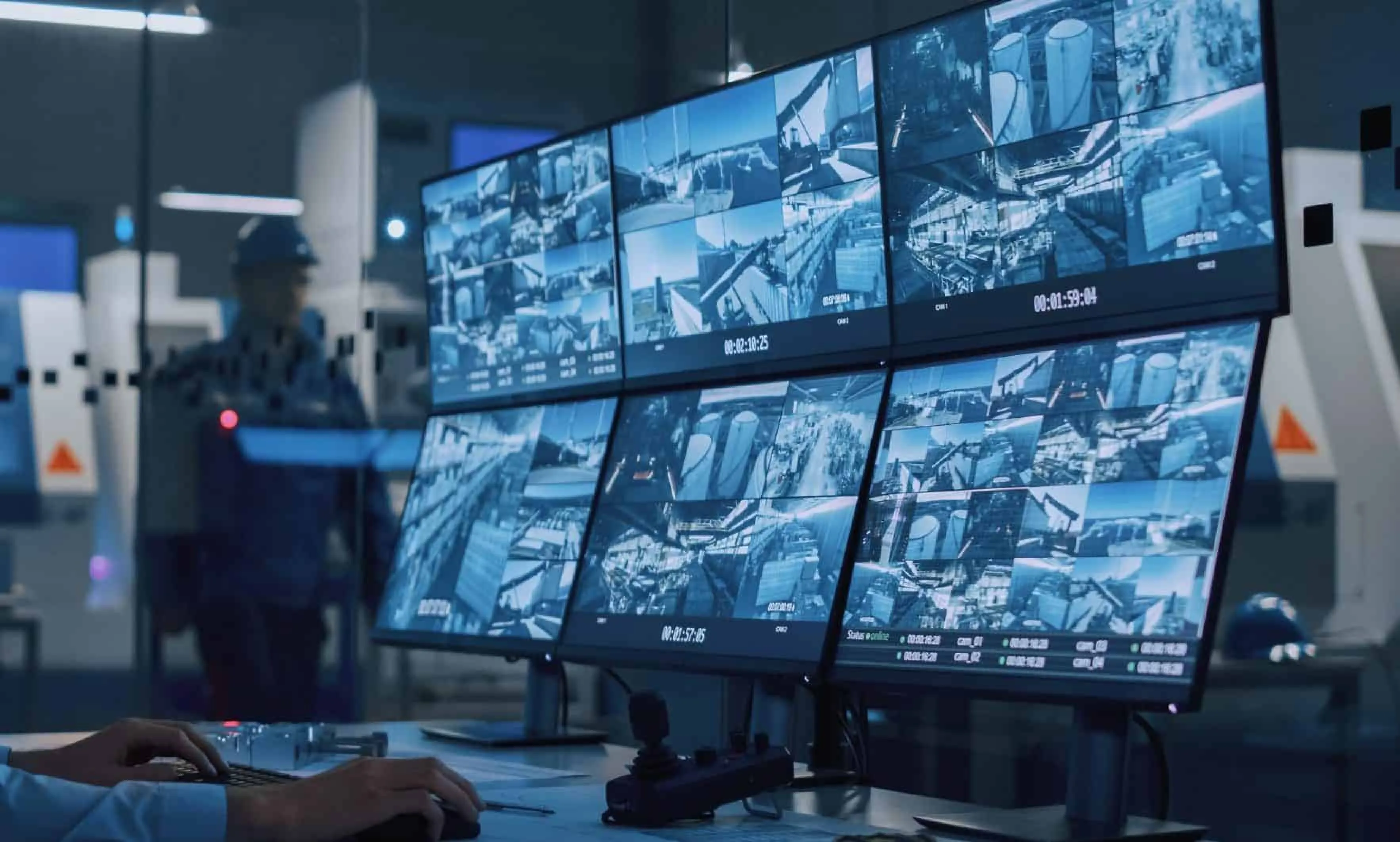 Multiple surveillance monitors displaying industrial facility camera feeds with a person working at the control desk.
