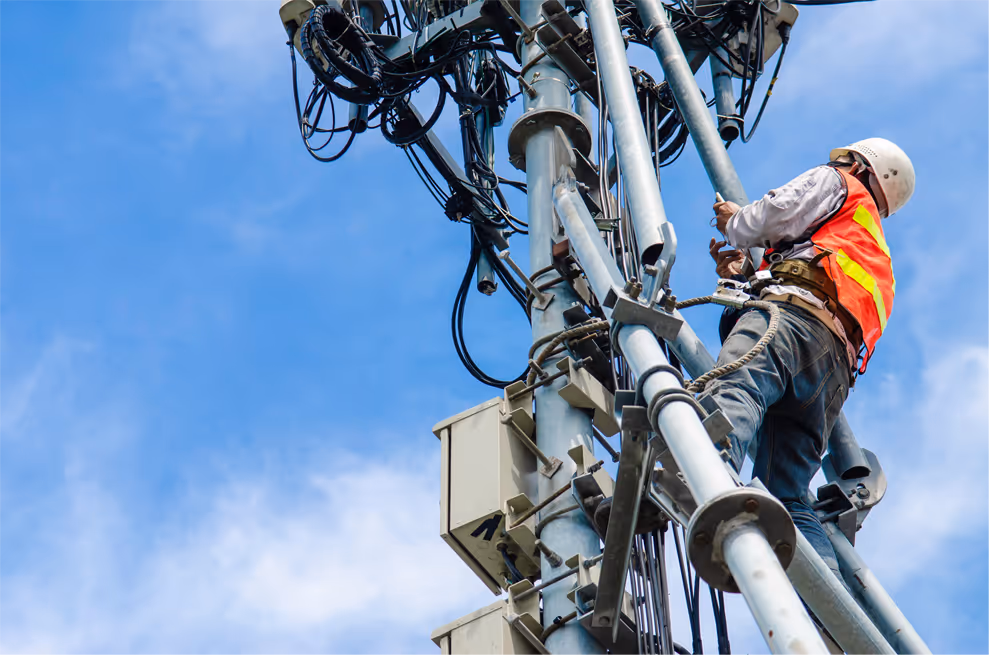 Wireless technician wearing a safety helmet and vest climbing a telecom tower with cables and equipment.